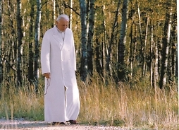Święty Jan Paweł II spacerujący drogą, przy której rosną drzewa. W ręku trzyma różaniec.