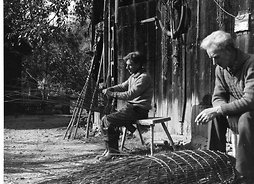 Zdjęcie archiwalne przedstawiające dwóch mężczyzn siedzących przed drewnianym budynkiem gospodarczym wykonujących przedmioty z wikliny