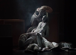 Scena ze spektaklu przedstawia siedzącą postać opartą o ścianę, a przy niej inne postaci leżące i siedzące powyginane w różnnych pozach..