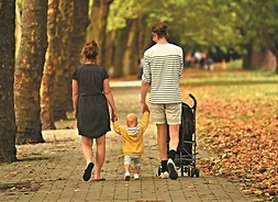 Mężczyzna i kobieta idą parkową aleją trzymając za ręce małe dziecko. Mężczyzna prowadzi wózek dziecięcy. Widok od tyłu.