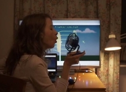 Kobieta siedzi bokiem przy biurku, na którym stoi włączony monitor komputera. Dmucha na elektryczny niewielki wiatraczek, który trzyma w dłoni.