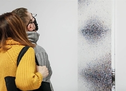 Dwie osoby oglądają abstrakcyjny obraz zawieszony na ścianie. Zdjęcie wykonane z boku.